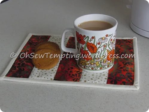 A white mug with a colorful floral design filled with tea or coffee sits on a patchwork fabric coaster, inspired by Quilt Patterns, next to two cookies on a light-colored countertop.