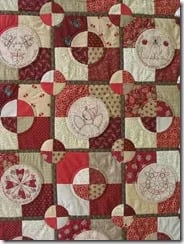 A patchwork quilt featuring red, cream, and green fabric squares arranged in a geometric pattern, with some panels displaying embroidered flowers and birds&mdash;perfect inspiration for exploring new quilt patterns.