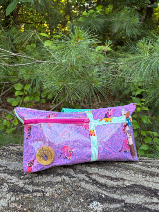 A purple, paisley-patterned Notion Bag with animal prints, colorful zippers, and decorative charms sits on a log, surrounded by green pine needles—perfect for carrying your sewing essentials in style.