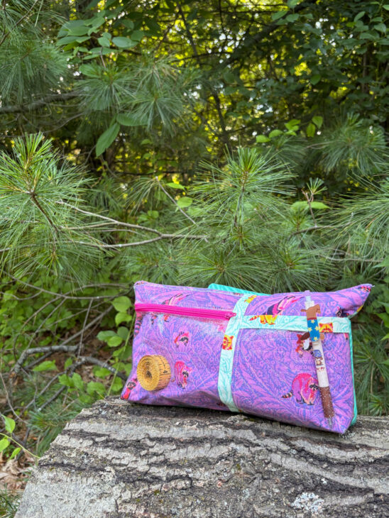 A vibrant notion bag with a purple pattern and pink-blue accents sits on a tree stump among green pine branches. A small wooden sewing item and a decorated stick dangle from the bag, set against lush greenery in the background.