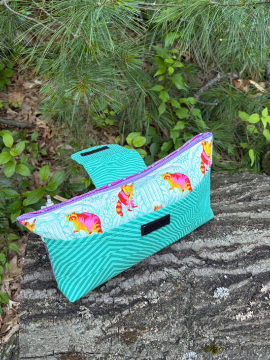 A colorful fabric pouch, crafted from a Sewing Bag pattern and featuring a cat motif with turquoise geometric design, sits on a tree stump amid green foliage and pine needles.