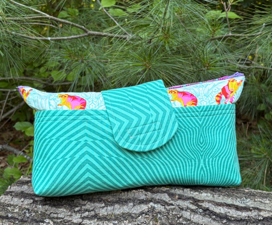 A turquoise quilted fabric Notion Bag clutch with diagonal stripe patterns and a flap closure, lined with a colorful floral print, rests on a tree log with pine branches in the background.