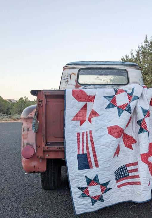 Sweet Liberty Quilt Along 4 A weathered red pickup truck is parked on a road, its tailgate down, with a Sweet Liberty Quilt Along white quilt decorated in red, blue, and patriotic patterns draped over the back. Trees and clear sky are in the background.