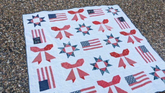 The Sweet Liberty Quilt features a white background with red, white, and blue patterns—bows, stars, and American flags—beautifully displayed on a bed of small stones outdoors.