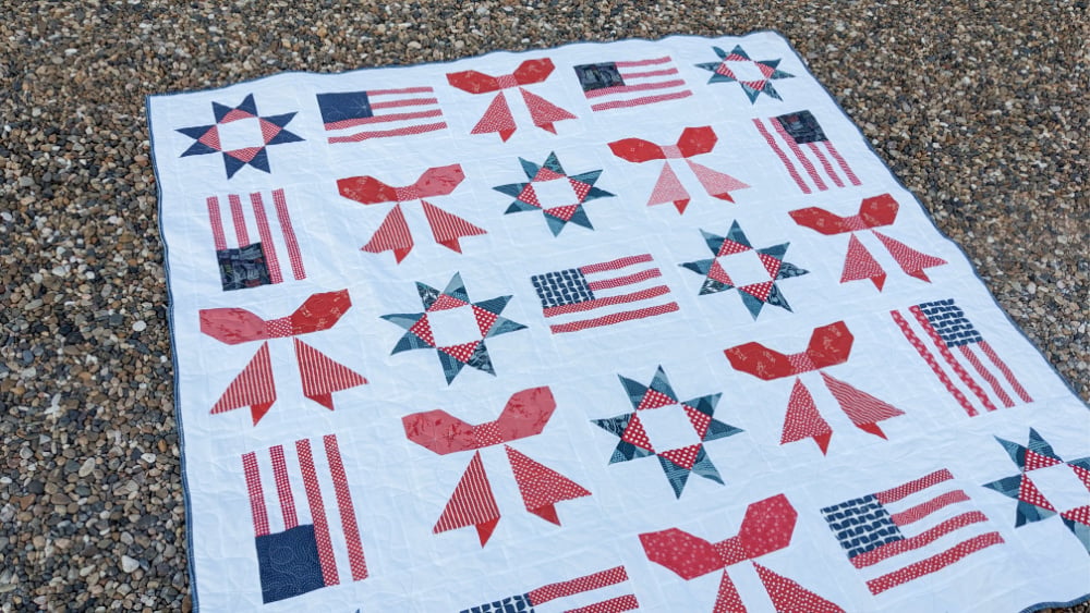 Sweet Liberty Quilt Along 3 The Sweet Liberty Quilt features a white background with red, white, and blue patterns—bows, stars, and American flags—beautifully displayed on a bed of small stones outdoors.