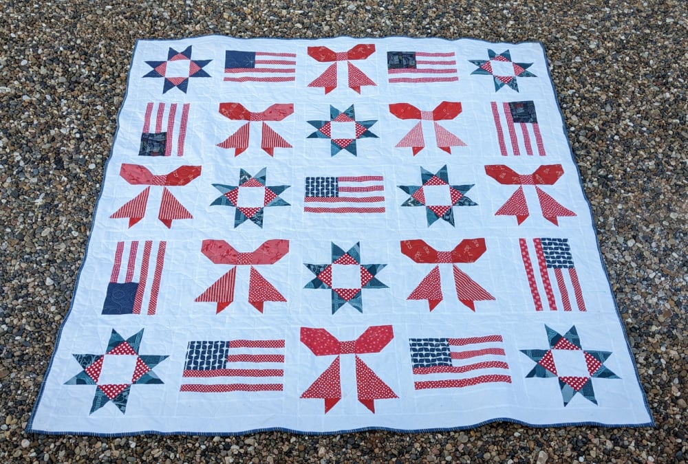 Sweet Liberty Quilt Along 5 A quilt with a white background featuring red, white, and blue patchwork designs—stars, American flags, and bows—lays on gravel, capturing the patriotic spirit of the Sweet Liberty Quilt Along.