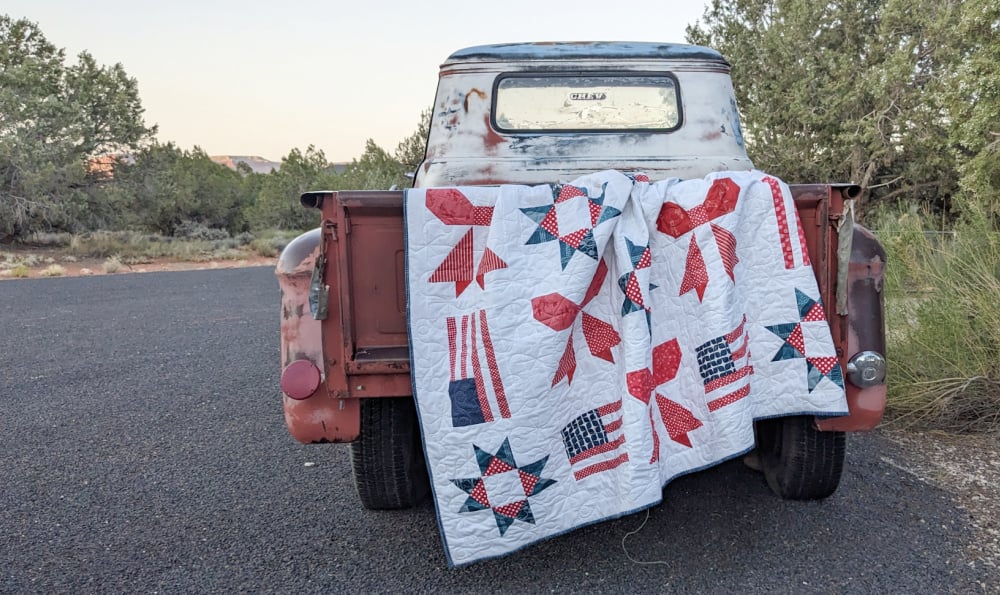 Sweet Liberty Quilt Along 2 A vintage, weathered pickup truck with a red, white, and blue patriotic Sweet Liberty quilt draped over its tailgate sits on a rural road surrounded by green trees—perfect inspiration for your next quilting or Quilt Along project.