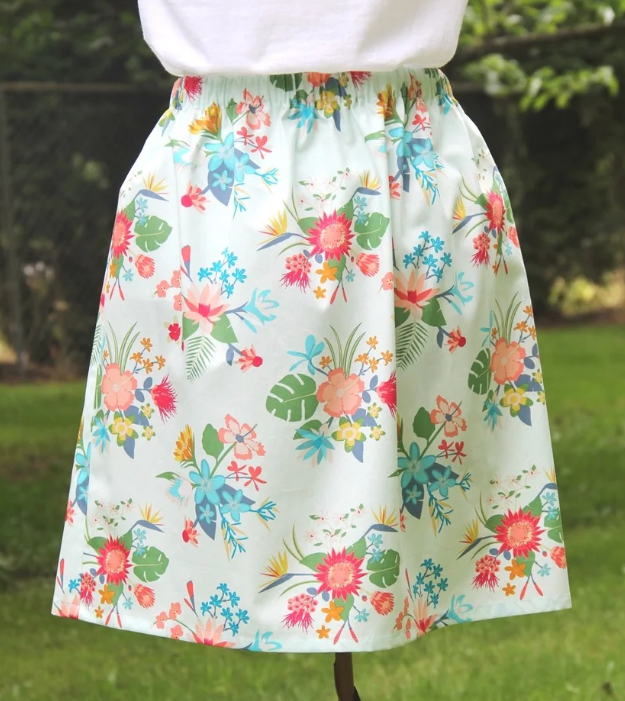 15 Easy Elastic Waist Skirt Patterns You Can Sew Today (All Free) 18 A person wearing a white shirt and a knee-length, light blue skirt with a colorful floral pattern—perfect inspiration for those looking for easy skirt patterns—stands outside on green grass near trees and a fence.
