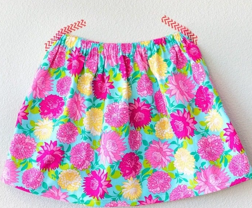 15 Easy Elastic Waist Skirt Patterns You Can Sew Today (All Free) 5 A colorful skirt with a floral pattern in pink, yellow, and purple on a blue background, pinned flat to a white wall with two pieces of red chevron-patterned tape—this charming piece is perfect for those looking for easy skirt sewing inspiration or searching for free skirt patterns.