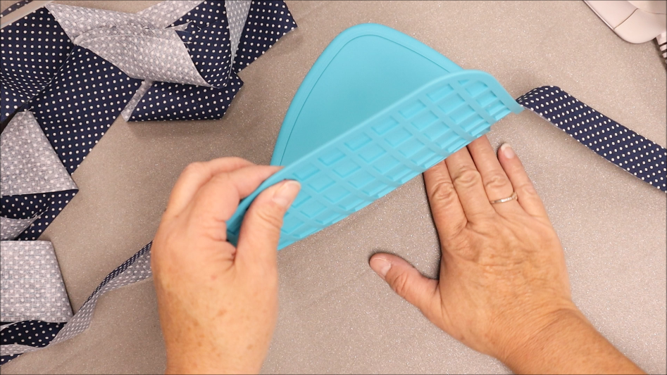 Two hands hold a blue silicone Binding Eaze ironing pad on a table, with navy and white polka-dotted fabric strips nearby—perfect for quilting projects from Quilted Heartz.