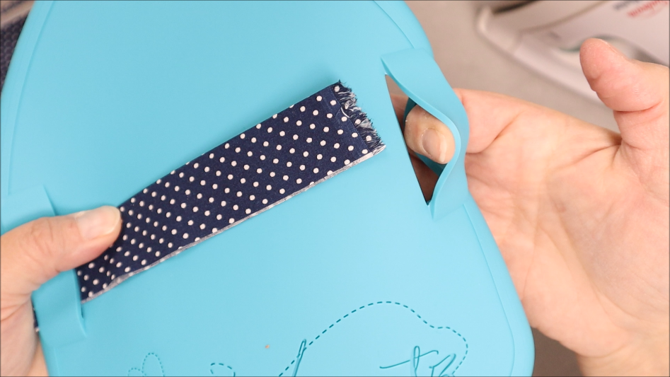 A hand threads a navy blue fabric strip with white polka dots through a slot in a blue silicone Binding Eaze ironing board, perfect for quilting projects. An iron is visible in the background, highlighting this essential Quilted Heartz tool in action.