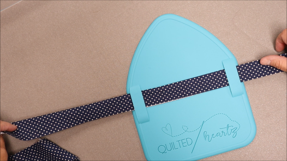 Hands pull a navy blue, polka-dotted fabric strip through the blue Binding Eaze quilting tool labeled "Quilted Heartz," making quilt binding easy and precise on a beige surface.
