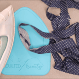 A hand holds an iron, pressing a polka-dot fabric strip on a blue ironing pad labeled "Quilted heartz," making quilting easier and neater with the help of Binding Eaze. More fabric strips are nearby on a gray surface.