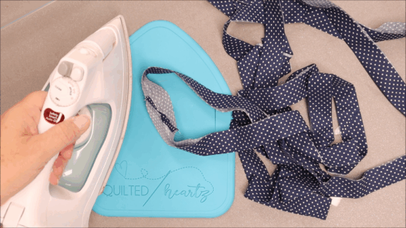 A hand holds an iron, pressing a polka-dot fabric strip on a blue ironing pad labeled "Quilted heartz," making quilting easier and neater with the help of Binding Eaze. More fabric strips are nearby on a gray surface.