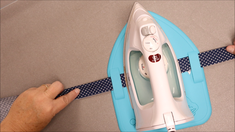 A person uses a white iron on a blue ironing mat to press a navy blue fabric strip with white polka dots, holding the strip straight with both hands—an essential step in quilting projects, especially when using Binding Eaze for perfect, crisp edges.