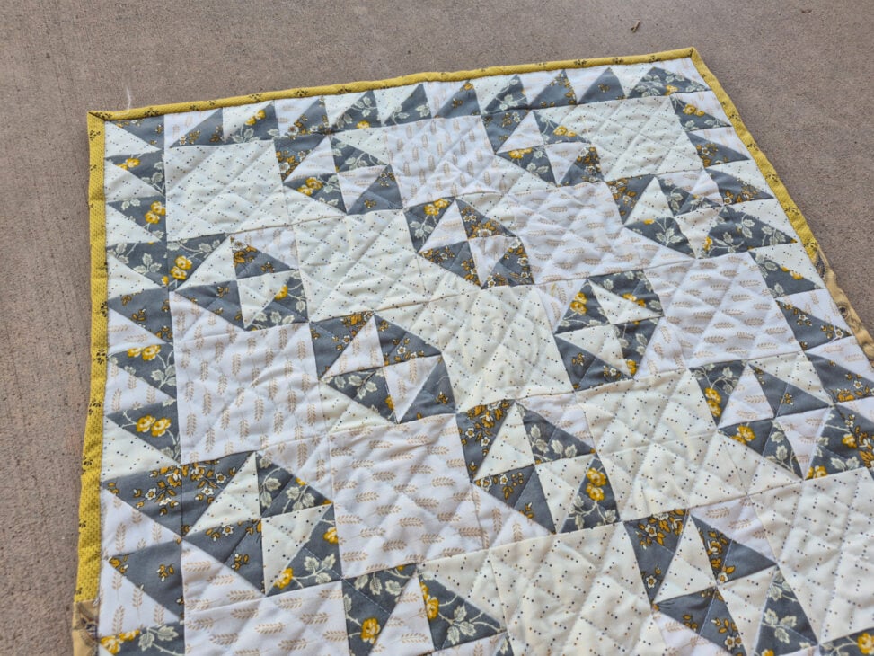 A handmade doll quilt features a geometric Broken Dishes pattern with gray, white, and yellow fabrics, including floral and dotted prints. The quilt is bordered with yellow fabric and laid flat on a concrete surface.
