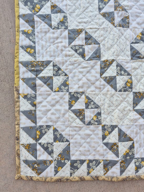 A close-up of a quilt corner with a patchwork Broken Dishes pattern in gray, white, and yellow fabrics, featuring floral and geometric prints, and a yellow fabric border on the edge.