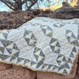 A gray, white, and yellow patchwork quilt featuring geometric Broken Dishes patterns is draped over a stone wall outdoors, with pinecones, rocks, and trees in the background.