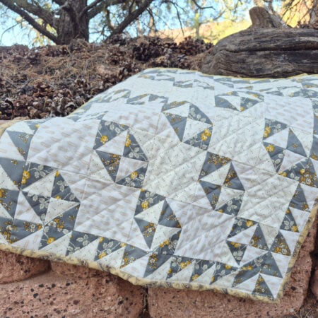 A gray, white, and yellow patchwork quilt featuring geometric Broken Dishes patterns is draped over a stone wall outdoors, with pinecones, rocks, and trees in the background.