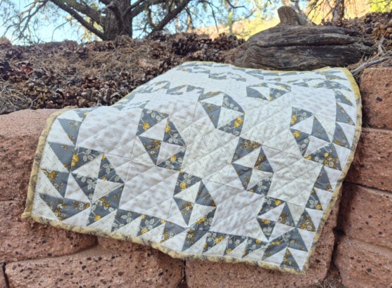 blog 3 A gray, white, and yellow patchwork quilt featuring geometric Broken Dishes patterns is draped over a stone wall outdoors, with pinecones, rocks, and trees in the background.