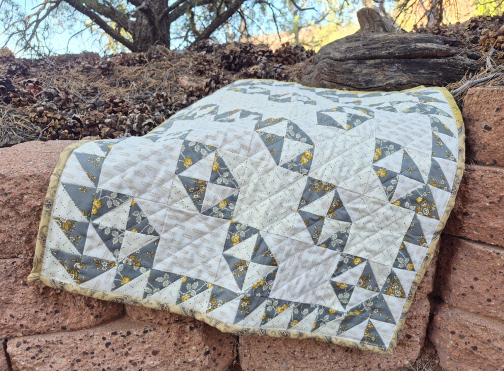 A gray, white, and yellow patchwork quilt featuring geometric Broken Dishes patterns is draped over a stone wall outdoors, with pinecones, rocks, and trees in the background.