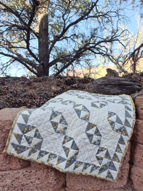 A gray and white Broken Dishes patterned quilt is draped over a stone wall outdoors, with dry pine needles, trees, and a blue sky in the background.