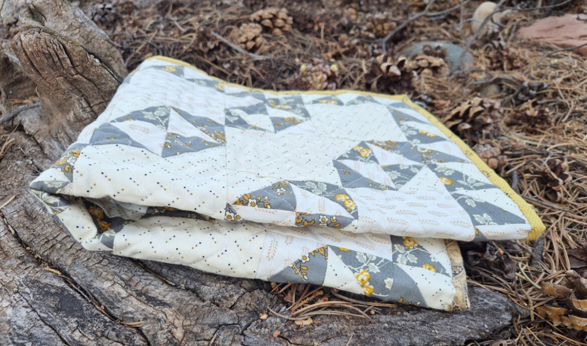 A folded patchwork Doll Quilt with gray, white, and yellow Broken Dishes patterns rests on a weathered tree stump surrounded by dry pine needles and pinecones.