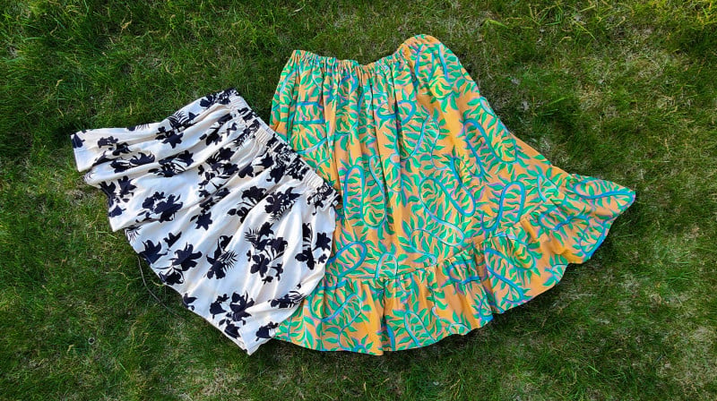 15 Easy Elastic Waist Skirt Patterns You Can Sew Today (All Free) 4 Two skirts lie on grass. One is white with black floral patterns, and the other is yellow with green and blue leafy patterns. Both have ruffled hems and gathered waists, perfect examples of easy skirt sewing or experimenting with elastic waist skirt patterns.
