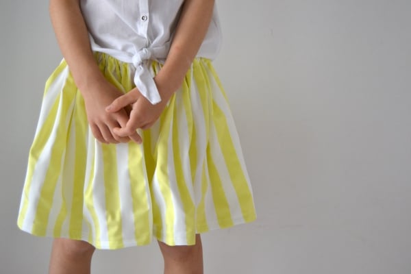 15 Easy Elastic Waist Skirt Patterns You Can Sew Today (All Free) 1 A child wearing a white shirt tied at the waist and a yellow-and-white striped skirt—perfect for those looking for easy skirt sewing inspiration—stands against a plain light background with hands clasped.