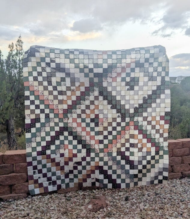 Free Online Creative Summit for Exploring New Hobbies 3 A handmade patchwork quilt with a geometric checkerboard pattern and various muted colors is displayed outdoors against a low brick wall and natural scenery, perfectly capturing the spirit of Exploring New Hobbies inspired by the upcoming Online Creative Summit.