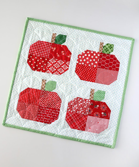 Mini Quilt Patterns You'll Want to Make This Weekend 17 A quilted square featuring four patchwork apples made from red patterned fabrics, each with a green leaf and brown stem, bordered by a light green edge on a white background with small blue dots—a charming mini quilt pattern.