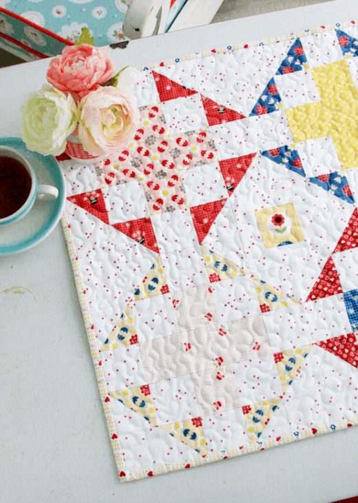Mini Quilt Patterns You'll Want to Make This Weekend 18 A colorful quilt with vibrant mini quilt patterns in red, blue, and yellow lies on a white table next to a blue cup of tea and a small vase holding pale pink and white flowers.