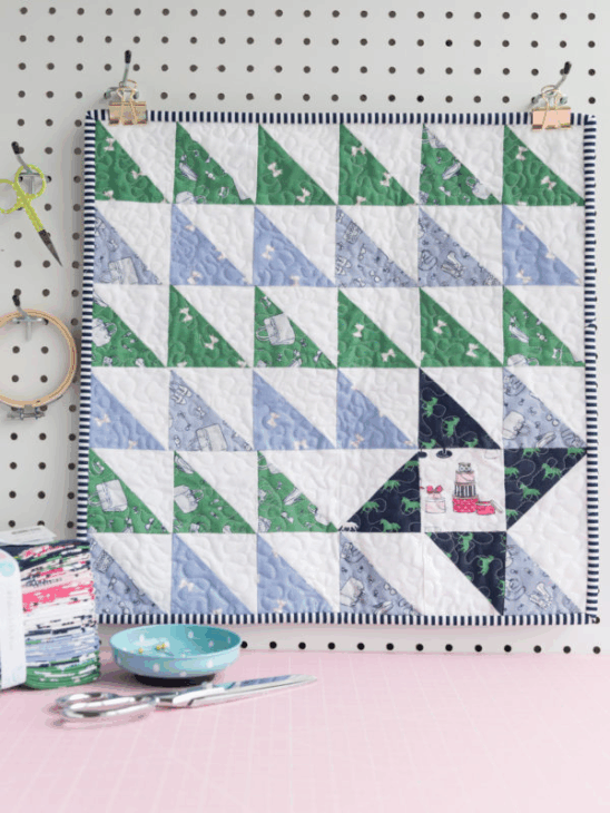 Mini Quilt Patterns You'll Want to Make This Weekend 19 A colorful quilt featuring green, blue, and white triangle quilt patterns hangs on a white pegboard wall, with sewing supplies like scissors, fabric, and a bowl placed nearby on a pink surface.