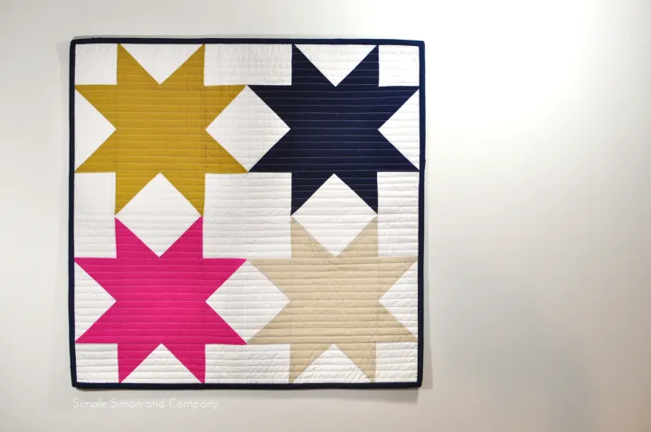 Mini Quilt Patterns You'll Want to Make This Weekend 20 A quilt with four large star patterns in mustard yellow, navy blue, hot pink, and beige on a white background, bordered with dark binding—an eye-catching design perfect for fans of modern quilt patterns—displayed on a plain white wall.