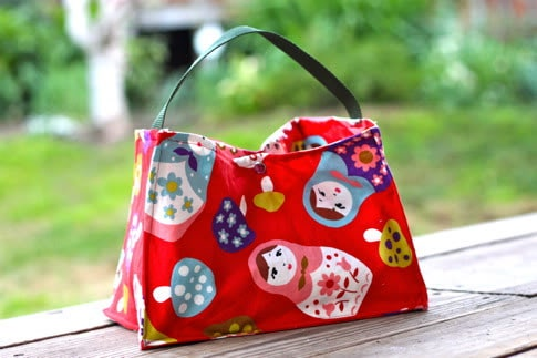 Easy and Cute Lunch Bag Sewing Patterns You'll Want to Make and Use 4 A bright red fabric bag with colorful matryoshka doll and mushroom patterns—perfect inspiration for cute lunch bag patterns—sits on a wooden surface outdoors, with grass and trees blurred in the background.