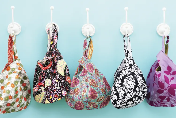 Easy and Cute Lunch Bag Sewing Patterns You'll Want to Make and Use 5 Five colorful patterned fabric bags hang on white hooks against a light blue wall. Showcasing cute lunch bag designs, these bags feature various floral and paisley patterns in different colors—perfect inspiration if you're searching for easy sewing patterns or unique lunch bag sewing patterns to craft your own stylish accessories.