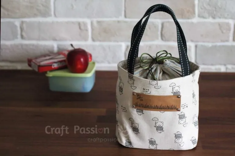 Easy and Cute Lunch Bag Sewing Patterns You'll Want to Make and Use 7 A small fabric lunch bag with black handles sits on a wooden table. Behind it are a green lunchbox, a red apple, and some snacks. The background features a white brick wall, making this scene perfect inspiration for anyone looking for cute lunch bags or searching for easy sewing patterns to create their own.
