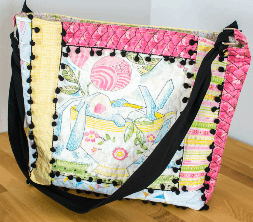 Quilt-As-You-Go Bag and Pouch Patterns You'll Want to Make 9 A colorful, quilted Quilt-As-You-Go fabric shoulder bag with floral and striped pouch patterns, decorated with black pom-pom trim, sits on a wooden surface against a white wall.
