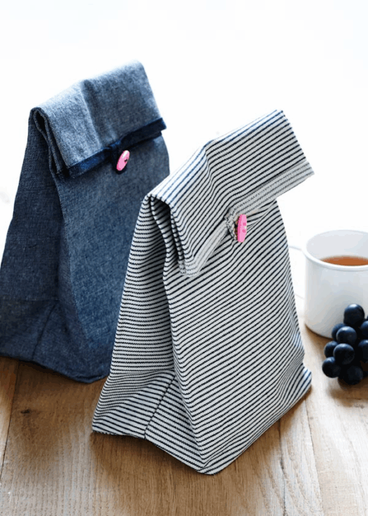 Easy and Cute Lunch Bag Sewing Patterns You'll Want to Make and Use 8 Two cute lunch bags—one denim and one striped—featuring pink buttons, sit on a wooden surface next to a white cup of tea and a small bunch of grapes. These charming accessories are perfect for those searching for easy lunch bag sewing patterns to create their own reusable fabric bags.