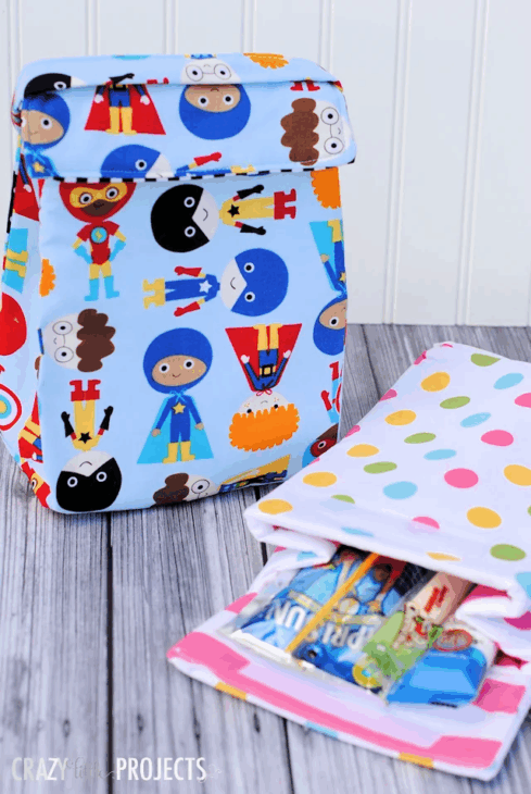 Easy and Cute Lunch Bag Sewing Patterns You'll Want to Make and Use 10 A colorful superhero-themed cute lunch bag, a polka-dotted napkin, and a zippered bag with snacks sit on a wooden surface. The lunch bag features cartoon superheroes on a light blue background, making it perfect inspiration for lunch bag sewing patterns.