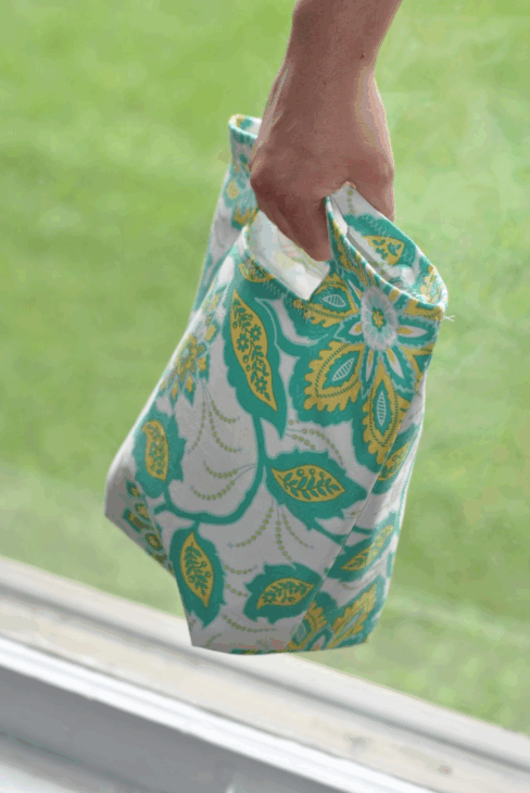 Easy and Cute Lunch Bag Sewing Patterns You'll Want to Make and Use 13 A hand carries a fabric lunch bag with a blue, green, and yellow floral pattern, showcasing one of the cute lunch bag patterns perfect for everyday use. The background is blurred green grass and a white window frame.