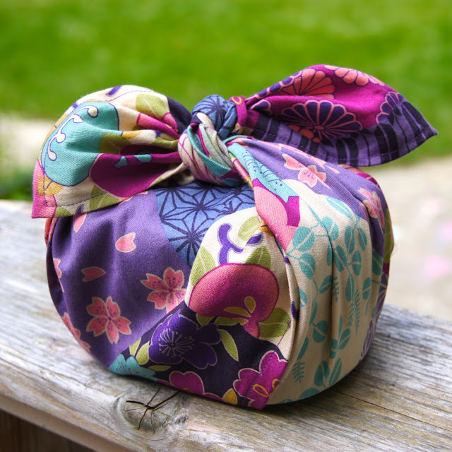 Easy and Cute Lunch Bag Sewing Patterns You'll Want to Make and Use 15 A colorful furoshiki cloth wraps an item with a knot on top, featuring floral patterns in purple, green, pink, and cream, placed on a wooden surface outdoors—an inspiring idea for those looking for cute lunch bags or exploring unique lunch bag sewing patterns.
