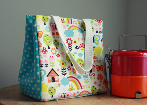 Easy and Cute Lunch Bag Sewing Patterns You'll Want to Make and Use 17 A colorful lunch bag with playful patterns and white handles, crafted from cute lunch bag sewing patterns, sits next to a red and silver tiffin container on a wooden surface.