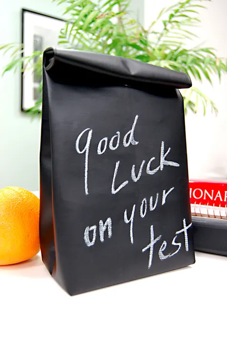 Easy and Cute Lunch Bag Sewing Patterns You'll Want to Make and Use 18 A black lunch bag with "Good Luck on your test" written in white chalk, sitting on a table next to an orange and some books, with a green plant in the background—just one example of the many cute lunch bags you can create using easy sewing patterns.