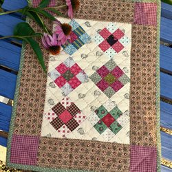 Mini Quilt Patterns You'll Want to Make This Weekend 6 A small, colorful patchwork quilt featuring Mini Quilt Patterns with floral and geometric designs, bordered by brown and purple fabric, is displayed on a blue wooden surface with pink coneflowers resting on top.