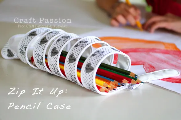 Pencil Pouch Sewing Patterns You'll Want to Make 16 A zippered, spiral pencil case holds colored pencils on a table. In the background, a child draws with crayons on white paper. Text reads, “Zip It Up: Pencil Pouch” and “Craft Passion – Free Sewing Patterns & Tutorials.”.