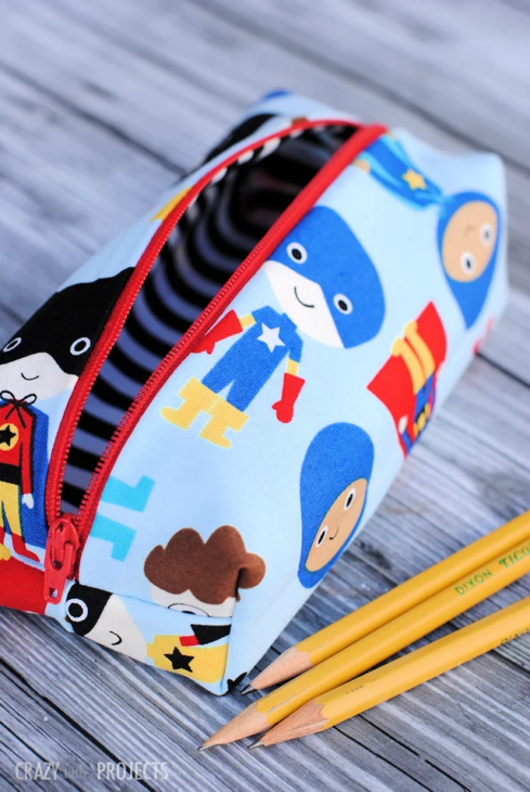 Pencil Pouch Sewing Patterns You'll Want to Make 17 A colorful pencil pouch made with vibrant sewing patterns featuring cartoon superheroes on the fabric, a red zipper, and three yellow pencils beside it, all arranged neatly on a wooden surface.