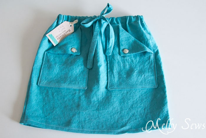 15 Easy Elastic Waist Skirt Patterns You Can Sew Today (All Free) 9 A turquoise skirt with a drawstring waist, front pockets with button flaps, and a clothing tag attached is laid flat on a white surface—perfect inspiration for those searching for easy skirt sewing ideas or free skirt patterns.