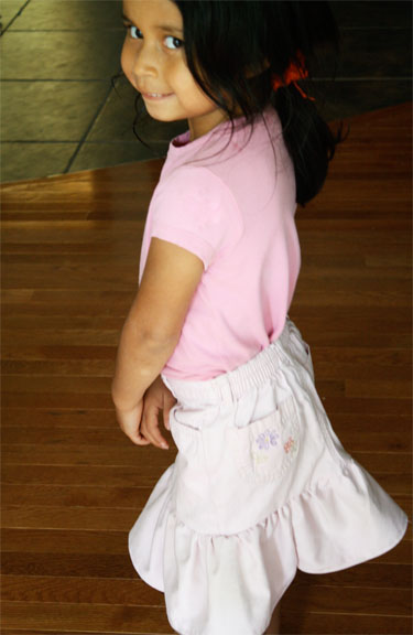 15 Easy Elastic Waist Skirt Patterns You Can Sew Today (All Free) 11 A young child with dark hair, wearing a light pink shirt and a skirt crafted from easy skirt sewing techniques, stands on a wooden floor and looks over her shoulder, smiling slightly at the camera.