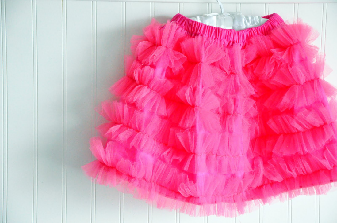 15 Easy Elastic Waist Skirt Patterns You Can Sew Today (All Free) 13 A bright pink ruffled tutu skirt with layers of tulle, inspired by easy skirt sewing techniques, hangs on a white hanger against a white paneled wall.